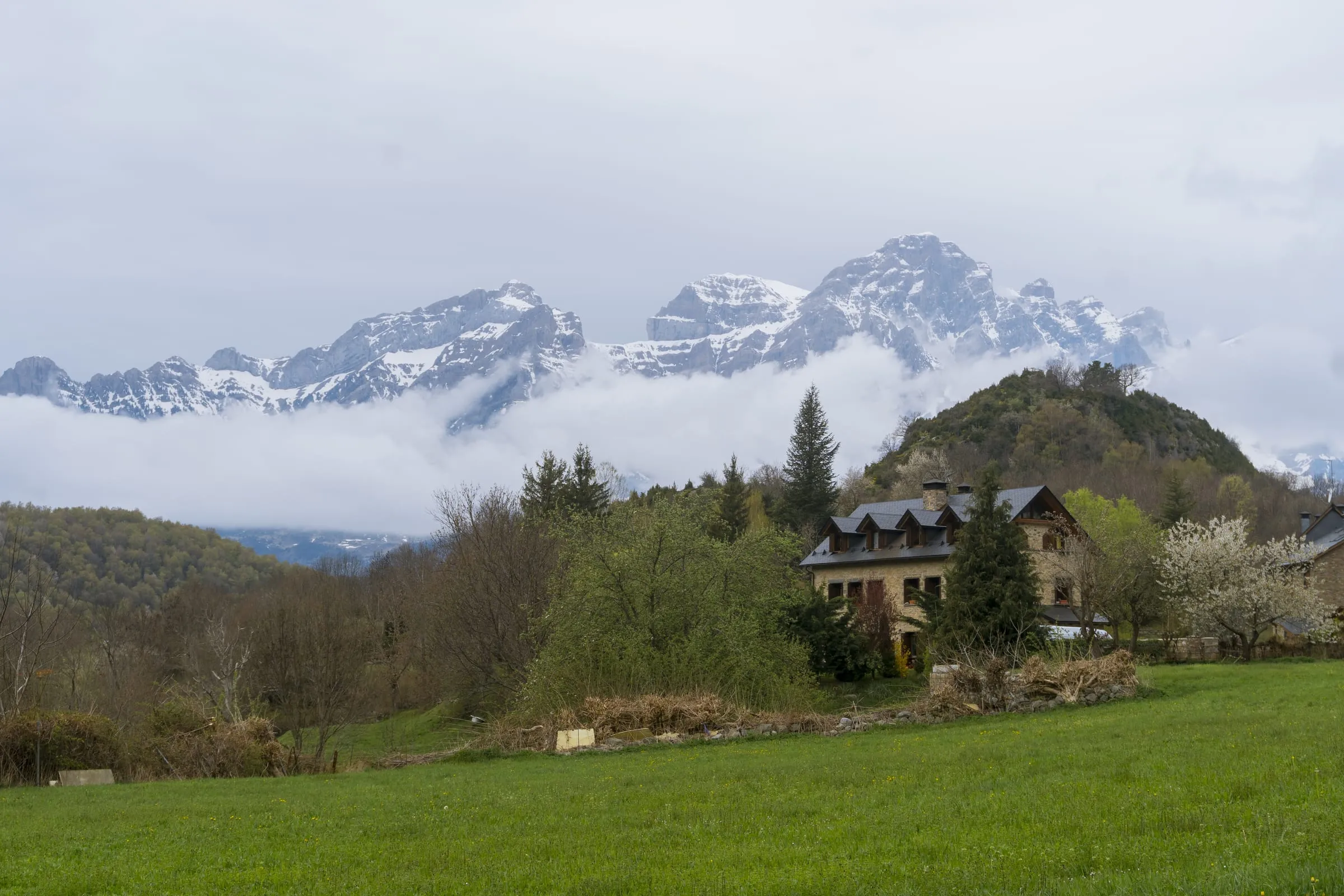 Casa de piedra en un valle verde con montañas nevadas y niebla al fondo